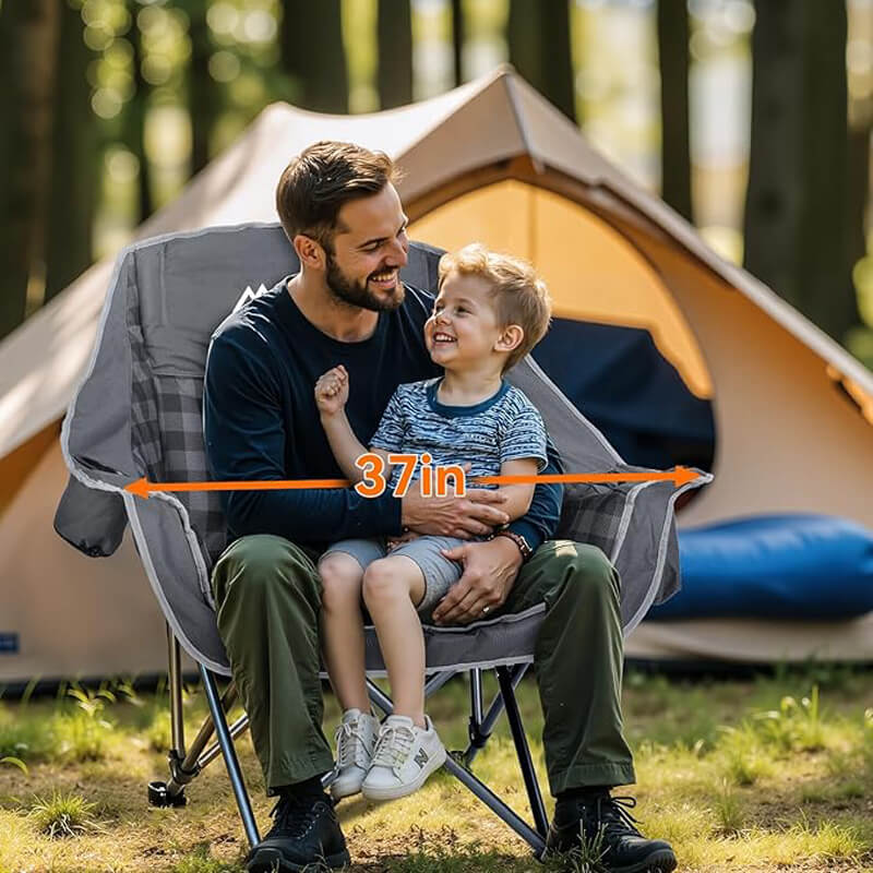 A father and young son sitting together on an oversized 37-inch wide folding camping chair with dark gray plaid padding, set against a tan camping tent in a sunlit forest campsite, ideal for family outdoor trips, camping, and picnics