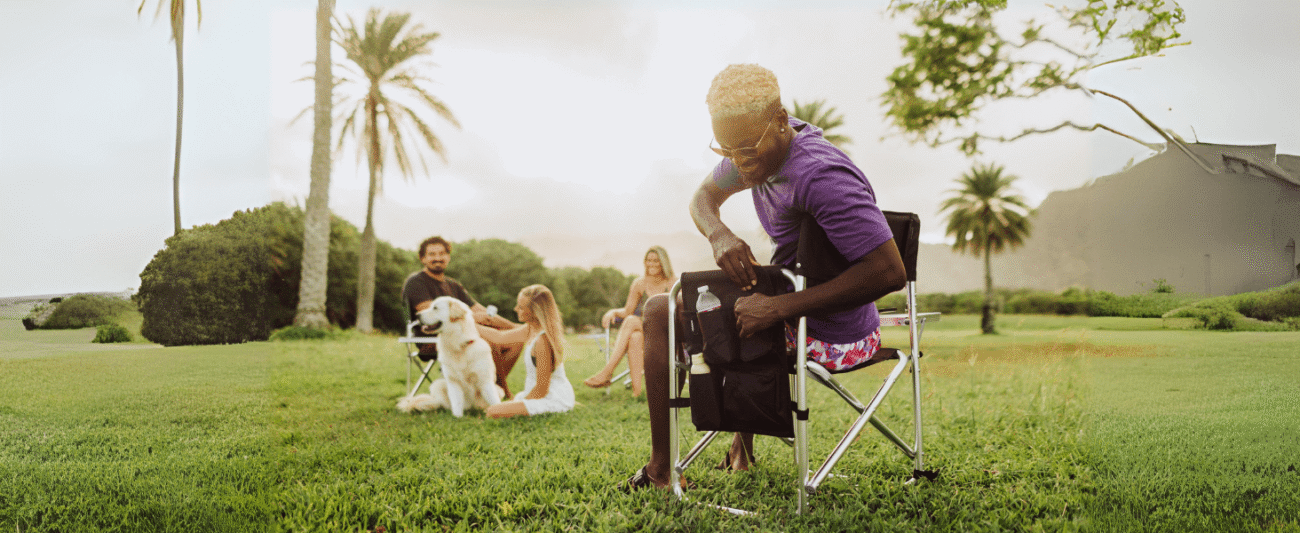 Man on a palm tree lawn using a director's folding chair with multiple pockets, showcasing storage functionality (for keys, phones, drinks, books, etc.