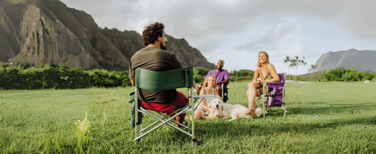 Multi-person tropical lawn leisure scene; display of multi-colored (green, purple) director's folding chairs; background of mountains and palm trees; interaction golden retriever; suitable for camping/picnicking/outdoor gathering scenarios; relaxed and comfortable atmosphere.