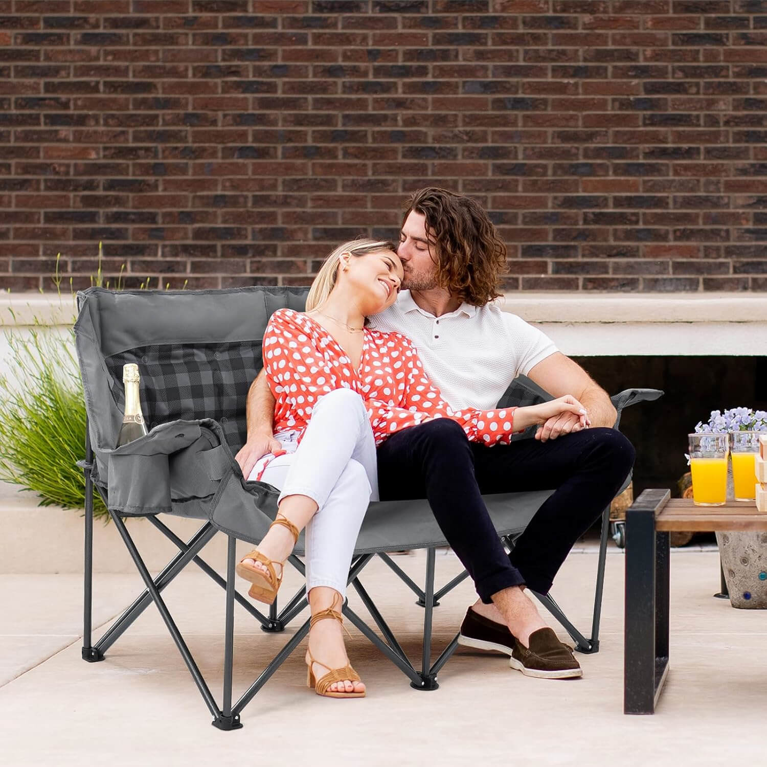 A cozy scene of a couple using gray double folding camping chairs on a terrace, featuring plaid-lined cushions and side storage pockets, comfortable and spacious, suitable for various outure settings.