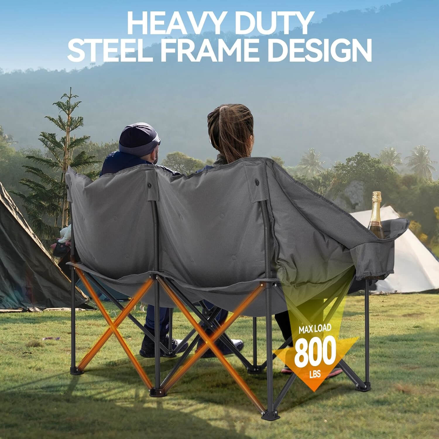 A gray double folding camping chair is placed on an outdoor lawn, with a background of green mountains, trees, and a camping tent. Two people sit comfortably leaningnst the backrest, admiring the scenery, with a bottle of champagne in the cup holder on the side, exuding a sense of leisure and relaxation.