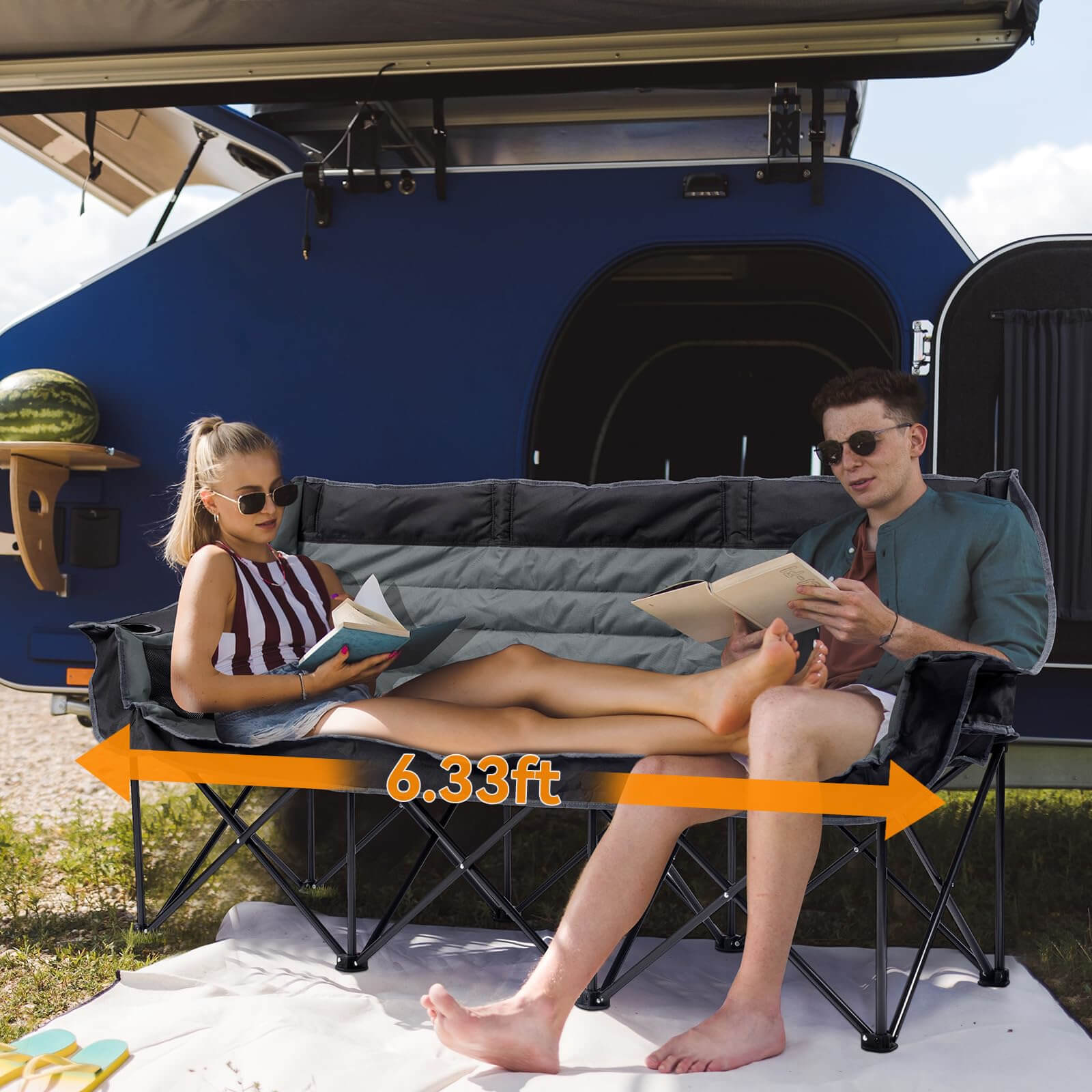 In an outdoor camping setting, a man and a woman sit barefoot on a black-and-grey two-tone folding three-seater sofa, reading. The sofa rests upon a white picnic mat, set against the backdrop of a blue RV. An orange double-headed arrow indicates the sofa's length of 6.33 feet, visually demonstrating its spacious dimensions and suitability for group relaxation.