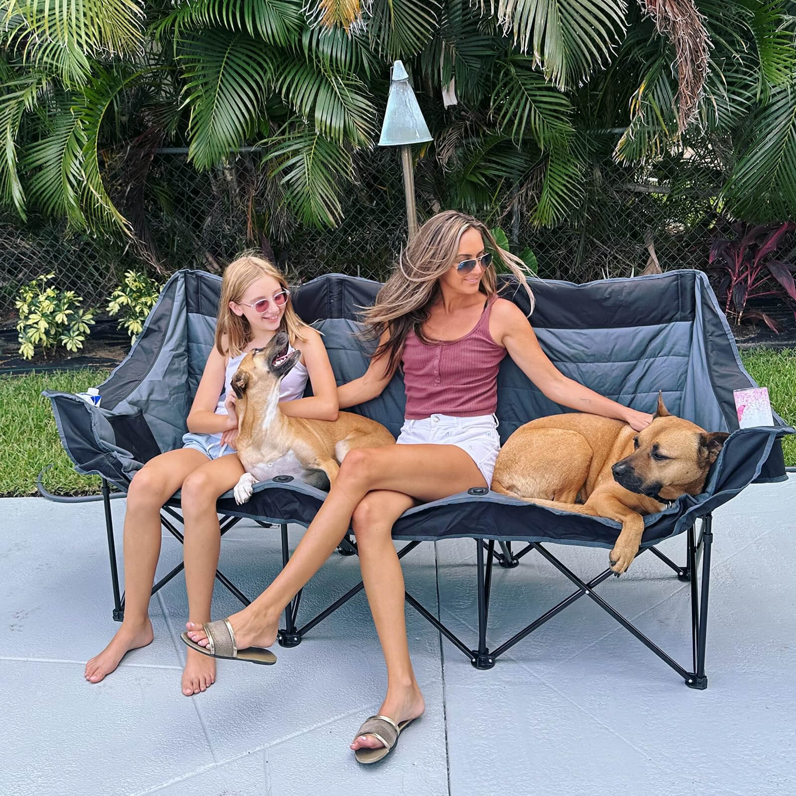In an outdoor patio setting, a woman and a young girl sit on a black-and-gray, color-block folding three-seater sofa, flanked by two large brown dogs snuggling close—creating a warm and cozy atmosphere. The sofa features built-in cup holders and storage pockets, while a backdrop of palm trees and fencing frames a scene of relaxed, casual family life outdoors.
