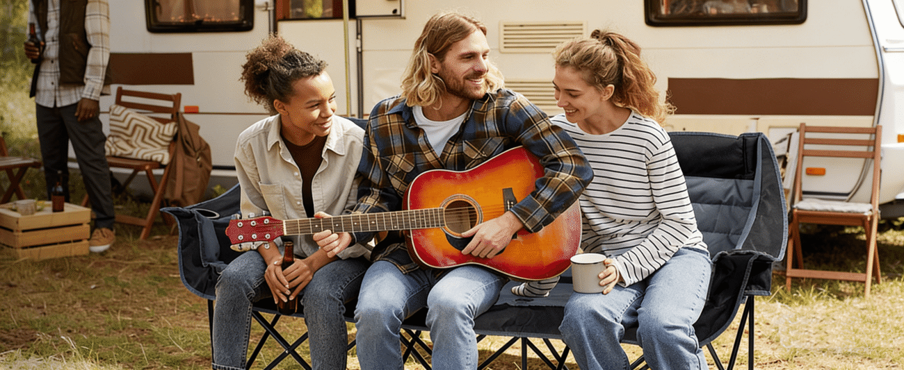 Three young people sit gathered in folding camping chairs in front of an RV. In the center, a long-haired young man cradles and plays a gradient-colored acoustic guitar; to his left, a curly-haired youth holds a beer bottle, while to his right, a young woman with a high ponytail clutches a mug. The trio interacts with smiles on their faces, set against a backdrop of an outdoor grassy area and camping furniture, creating a relaxed and pleasant atmosphere.