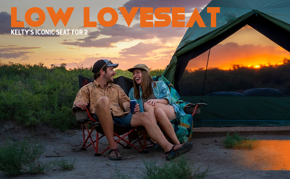 A cheerful couple relaxes side by side on Kelty Low Loveseat camping chairs during a sunset outdoor adventure. The man, wearing a cap and casual shirt, shares a laugh with the woman in a hat and open shirt, with a green tent set up against a vibrant orange and purple sky backdrop. Text overlay reads "LOW LOVESEAT" and "KELTY'S ICONIC SEAT FOR 2