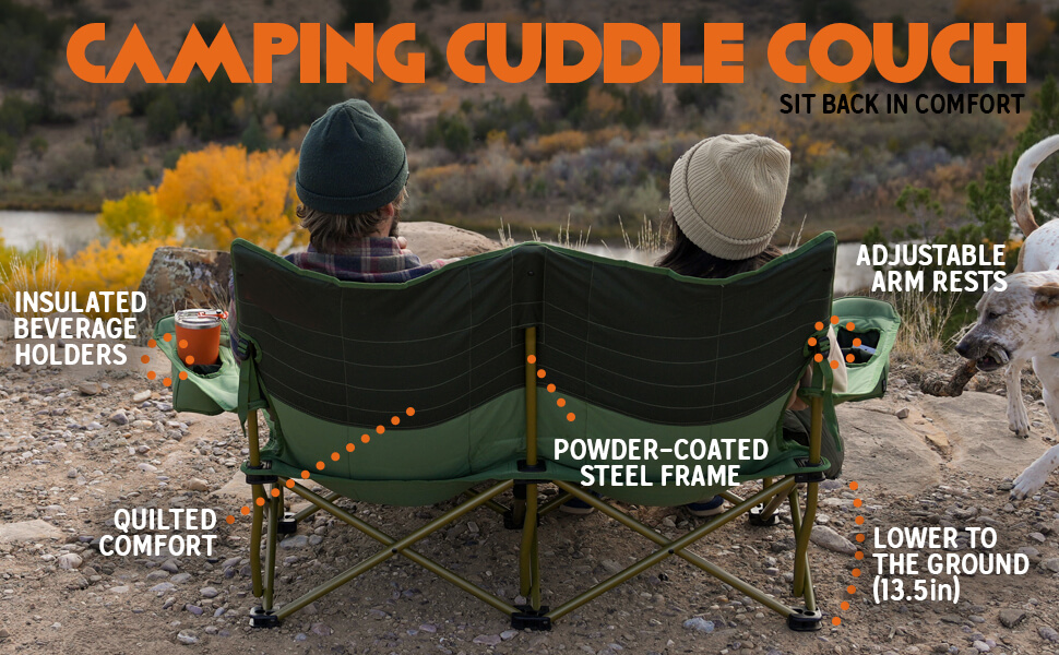 Back view of two people sitting together on a green "Camping Cuddle Couch" folding chair, overlooking a scenic autumn landscape with a river and trees. A white spotted dog stands nearby. Orange text at the top reads "CAMPING CUDDLE COUCH" and "SIT BACK IN COMFORT". Annotated features include insulated beverage holders, quilted comfort, powder-coated steel frame, adjustable arm rests, and a low-to-the-ground design (13.5in)