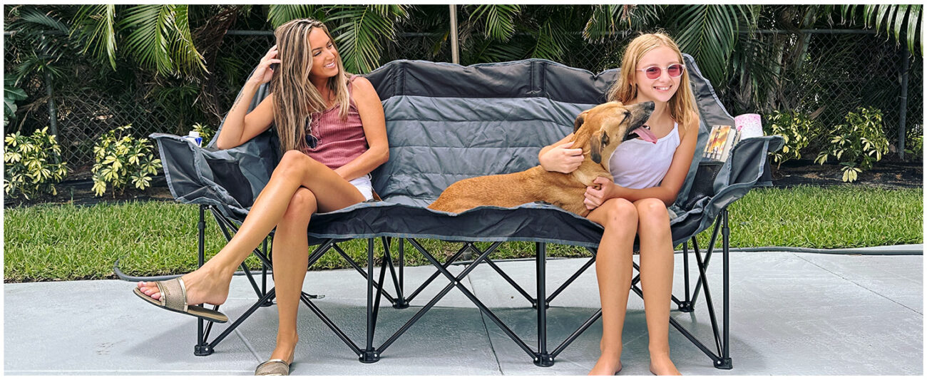In an outdoor patio setting, two women and a brown dog are seated on a tri-fold camping sofa featuring a two-tone gray and black design. The woman on the left, dressed in a sleeveless pink-purple top and white shorts, strikes a relaxed pose; the young woman on the right, wearing a white tank top and pink sunglasses, is cradling the dog—which has its tongue playfully lolling out. With items resting in the cup holders on both sides of the sofa, the scene exudes a warm and cozy atmosphere, effectively showcasing the product's suitability for family leisure in an outdoor setting.
