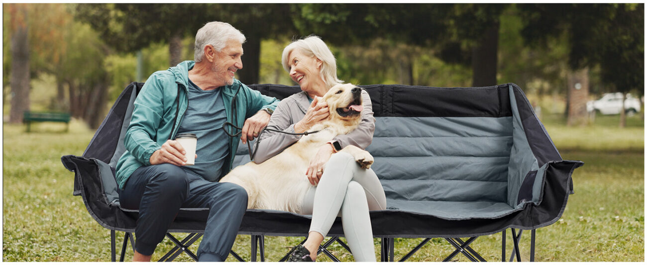 On the grassy lawn of a park, an elderly couple—both white-haired—sit upon a two-tone black-and-grey, three-seater folding camping sofa, with a Golden Retriever nestled affectionately by their side. The gentleman holds a cup of coffee, while the lady gently strokes the dog; as they exchange smiles, the warm and relaxed atmosphere beautifully illustrates how the product is perfectly suited for family outdoor leisure.
