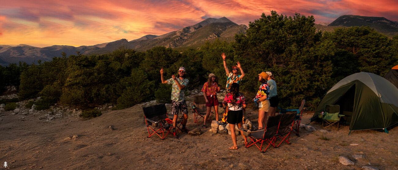 A wide panoramic shot of a group of seven friends celebrating around a campfire at a mountain campsite during a vibrant sunset. They stand on rocky dirt ground, surrounded by red-framed camping chairs, a green tent, and dense evergreen trees. The sky glows with orange and pink hues, with rugged mountain peaks visible in the background