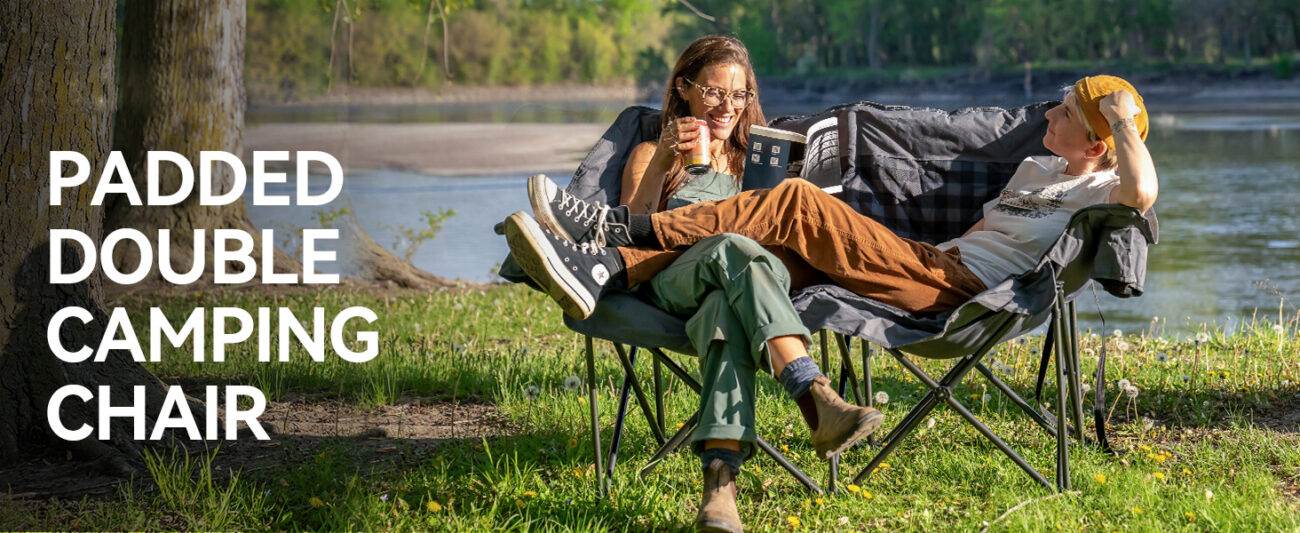 Padded double camping loveseat with two people relaxing by a river, holding drinks, showing the chair's side pockets and comfortable design.